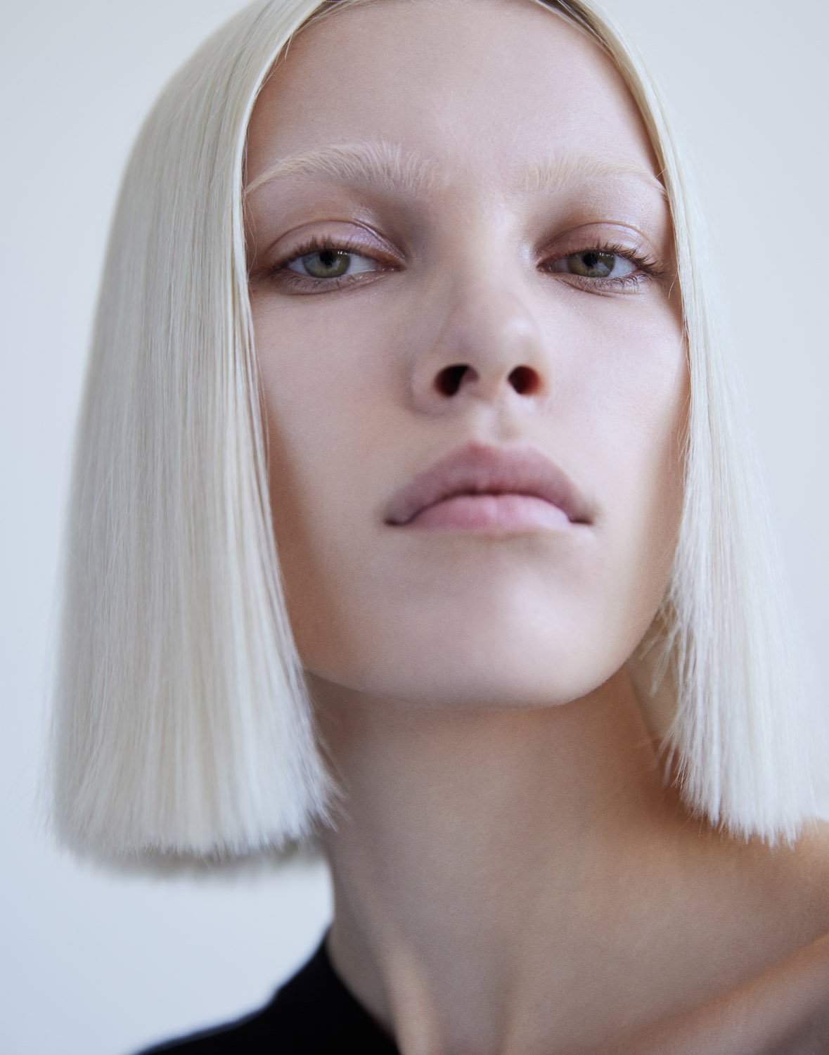 Close-up portrait of a woman with white blonde bob hair and bleached eyebrows against a plain white background